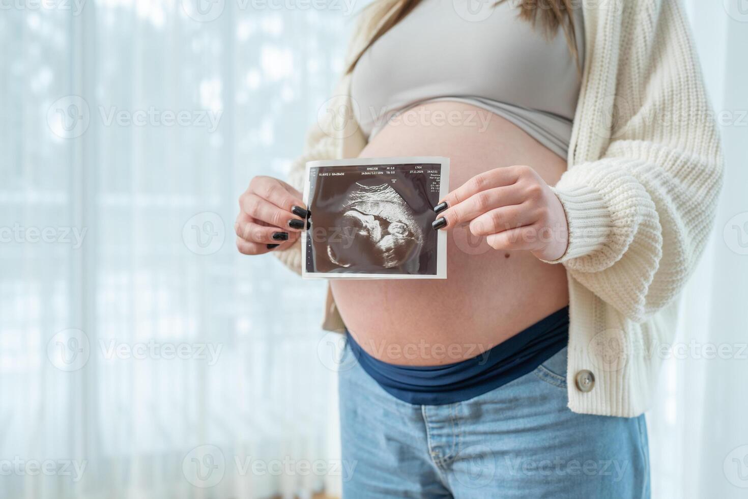 Young pregnant woman holding ultrasound scan photo of a fetus in front of her belly in third trimester at home with copy space. Concept of pregnancy, sonography. Maternity prenatal care