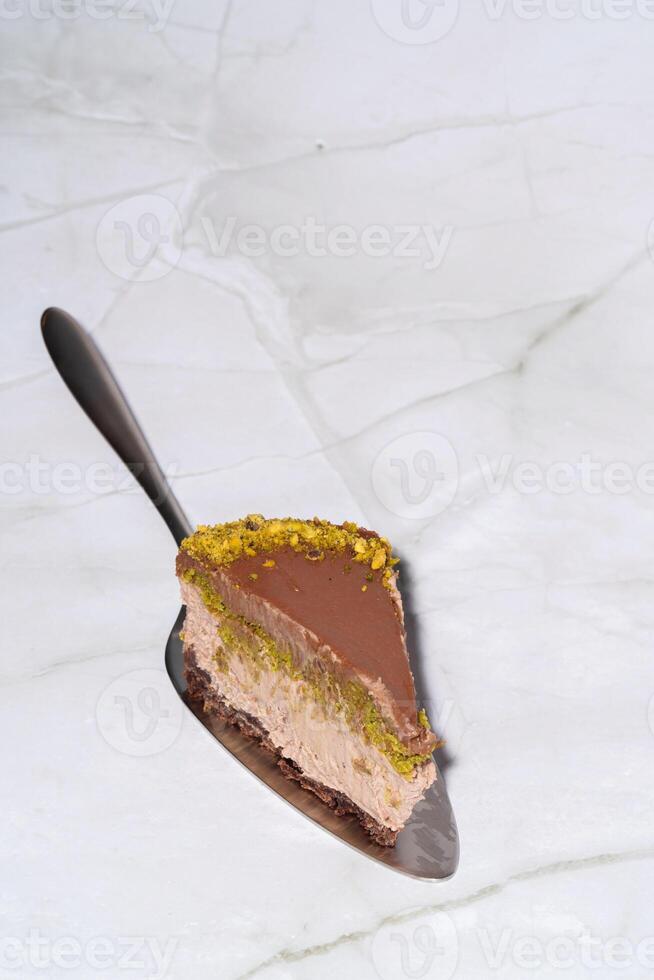 Slice of chocolate Dubai cheesecake with pistachio paste and kataifi covered in chocolate ganache on cake spatula. White background with copy space. Vertical photo. Indulgence, gourmet desserts. photo