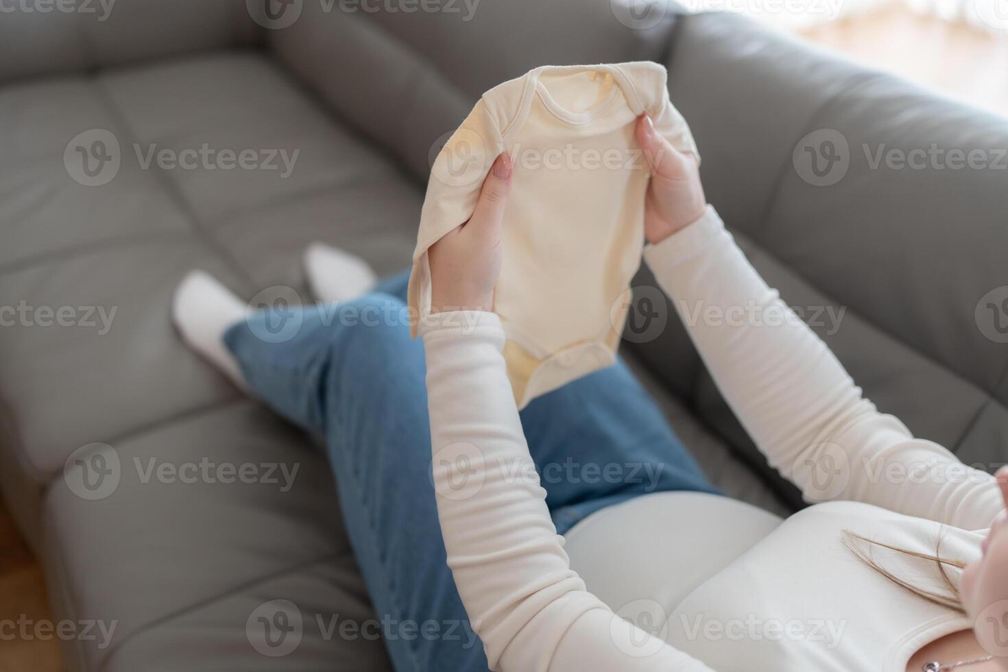 Pregnant woman holding baby onesie bodysuit sitting on a sofa. Preparing and planning baby clothes for new baby, packing for maternity at home photo