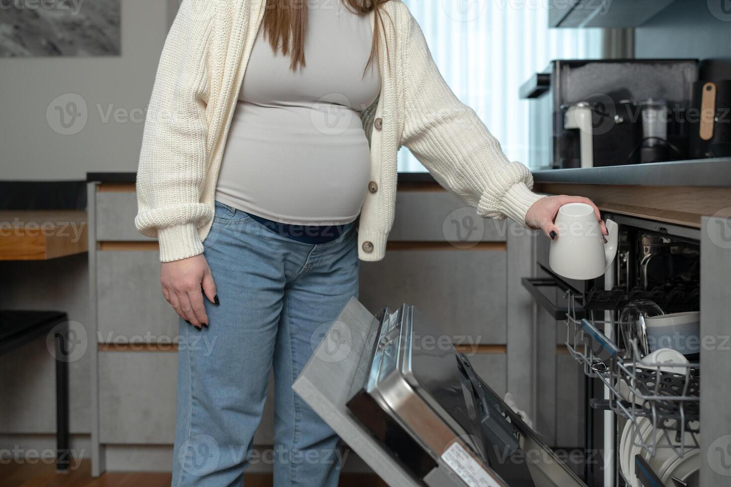 Close up of pregnant woman loading a dishwasher in the kitchen. Concept of daily life, home chores, and balancing household responsibilities during maternity. photo