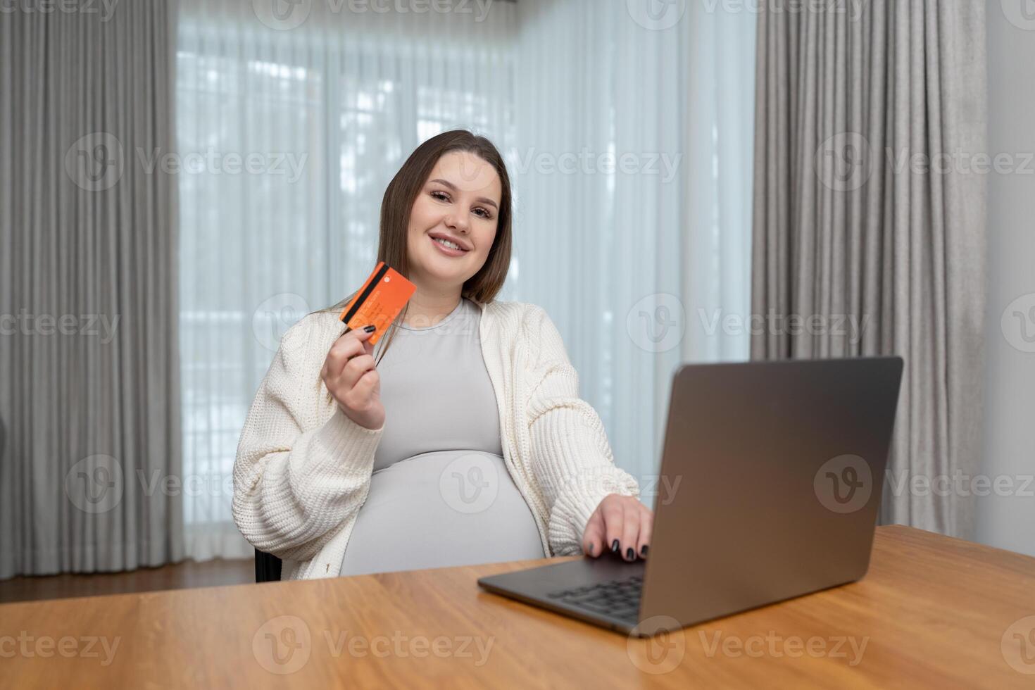 Smiling pregnant woman shopping online with a credit card and laptop at home with copy space looking at camera. Concept of convenience, ecommerce, and preparing for motherhood through online shopping. photo