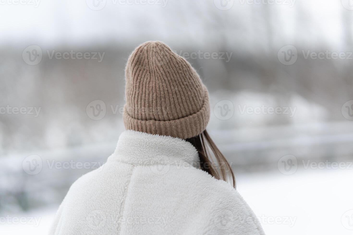 Woman in a winter beanie and white fluffy jacket during winter day having a walk outside, view from behind. Cozy winter outerwear, cold weather and peaceful connection with nature during snowy weather photo