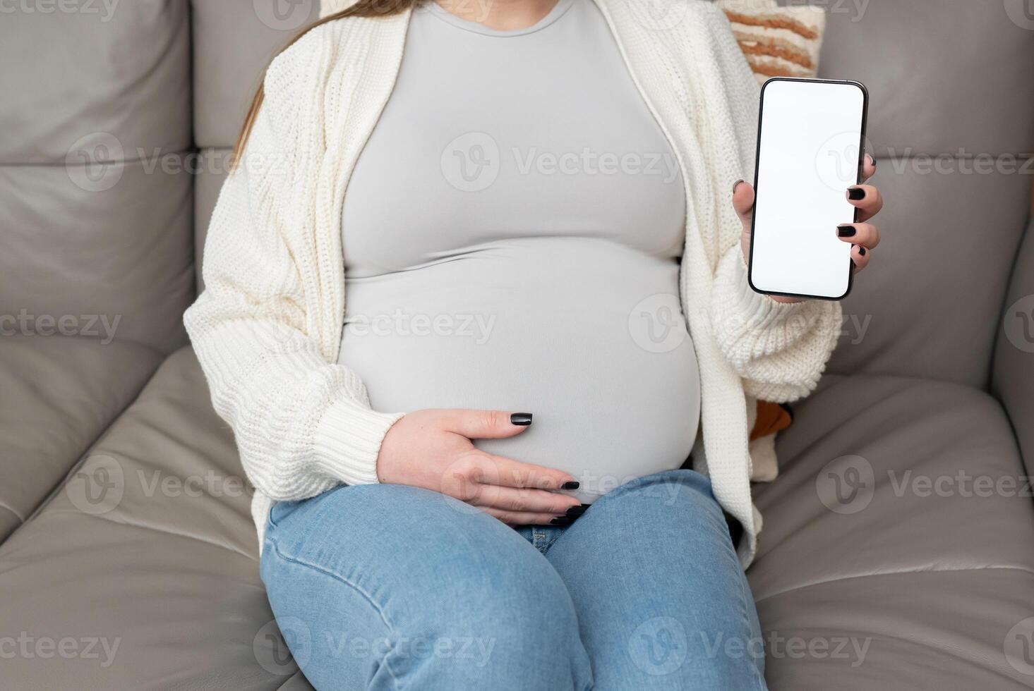 Close-up of pregnant woman holding and showing mobile phone with empty display at home. Mockup image with copy space for app or interface customization photo