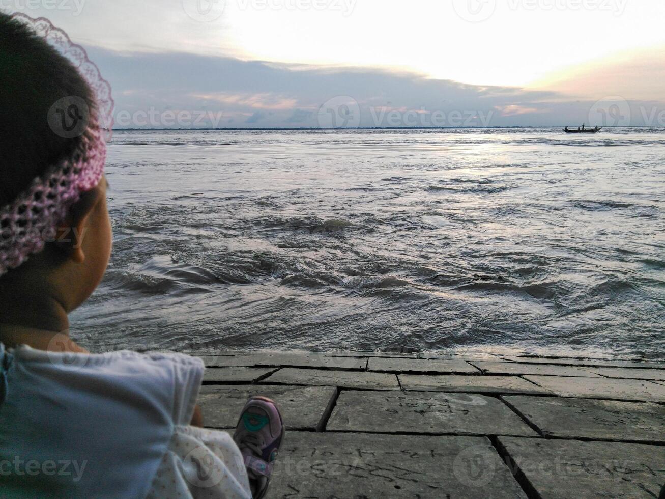 A serene sunset over a body of water with a person sitting on a paved area near the water. A boat with two people is visible on the horizon, and the partly cloudy sky adds to the tranquil atmosphere. photo