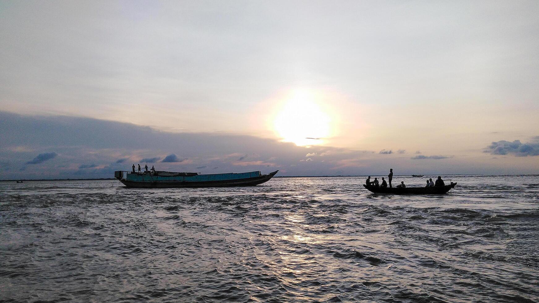 A serene sunset over a body of water. Two boats, one larger and one smaller, are visible. The partly cloudy sky and warm glow create a tranquil atmosphere. photo