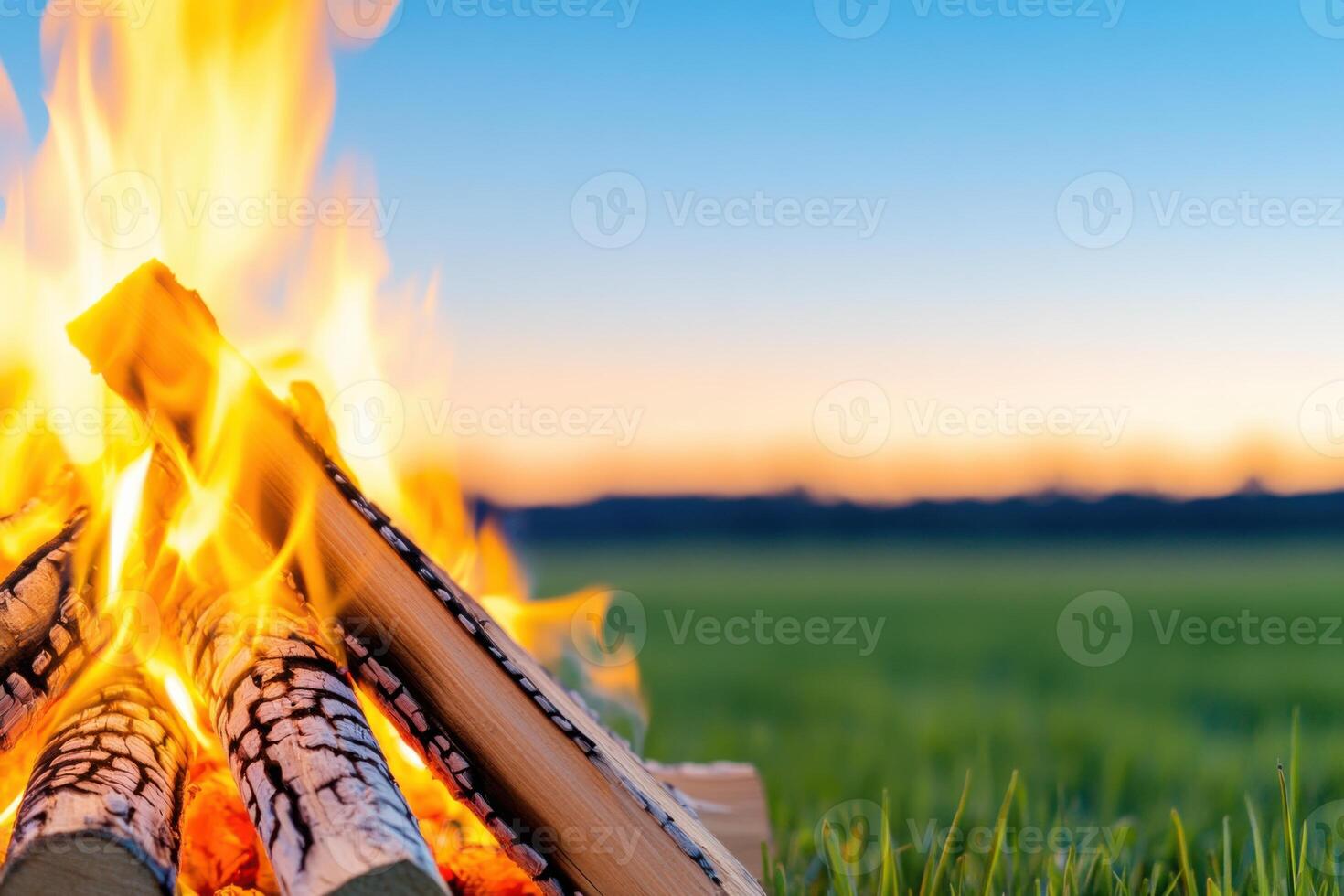 Campfire crackles with warmth as dusk settles over an open field with grass under a dusky blue sky and logs encircling the blazing fire photo