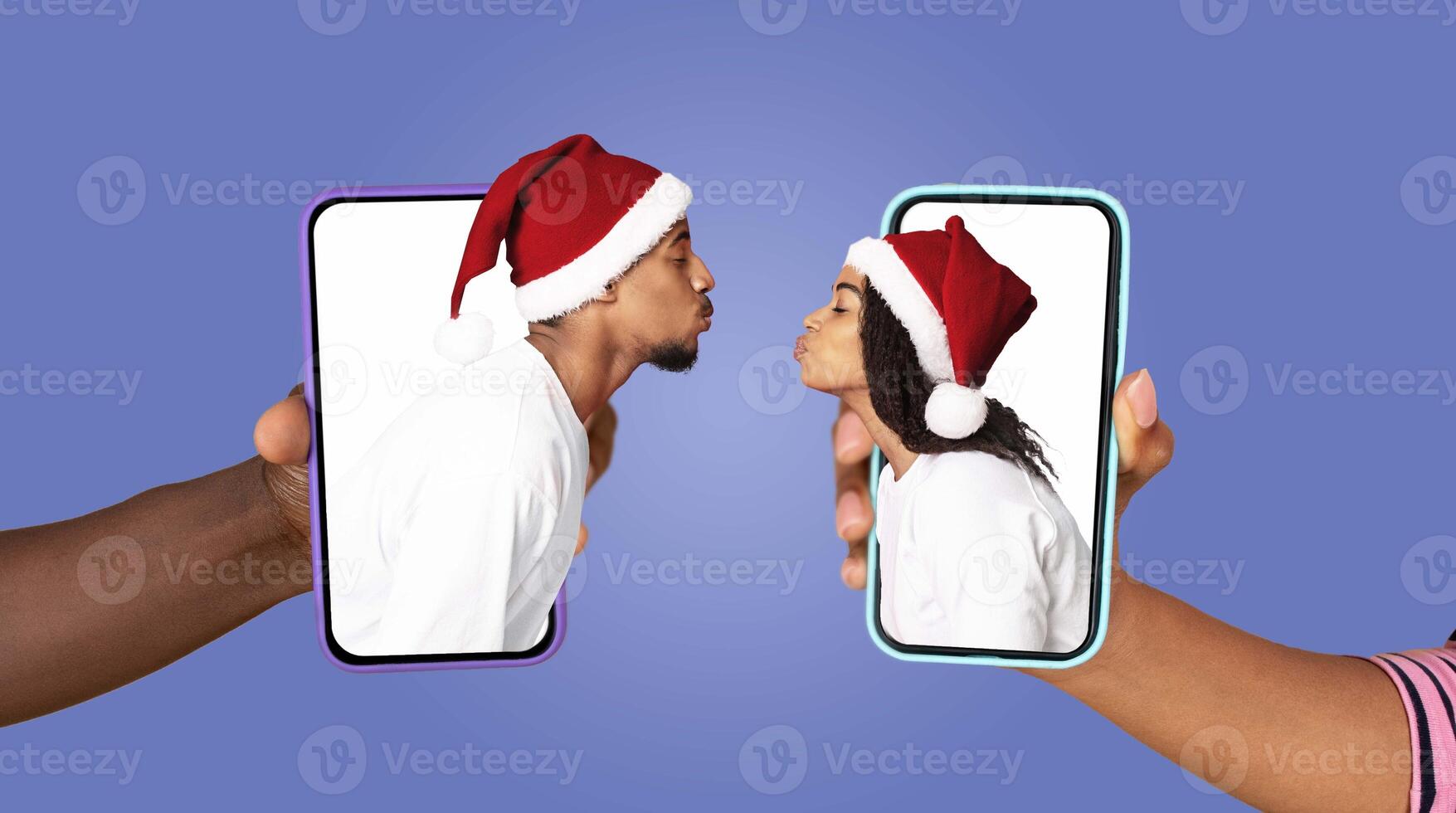 Two people in festive hats connect affectionately on their smartphones, sharing a kiss through the screens. They express love while embracing modern technology during the holiday season. photo