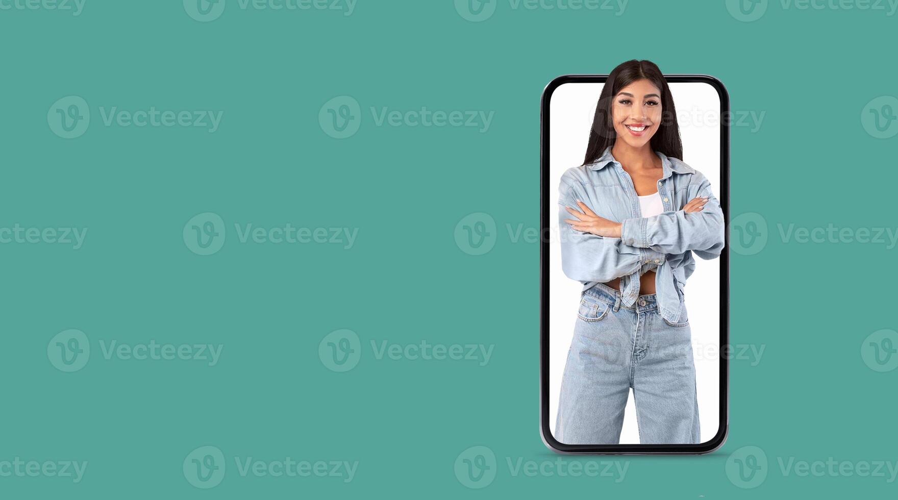 A young woman confidently poses with arms crossed, showcasing her cheerful demeanor on a large smartphone screen. The background is a solid color, emphasizing her presence and the technology. photo