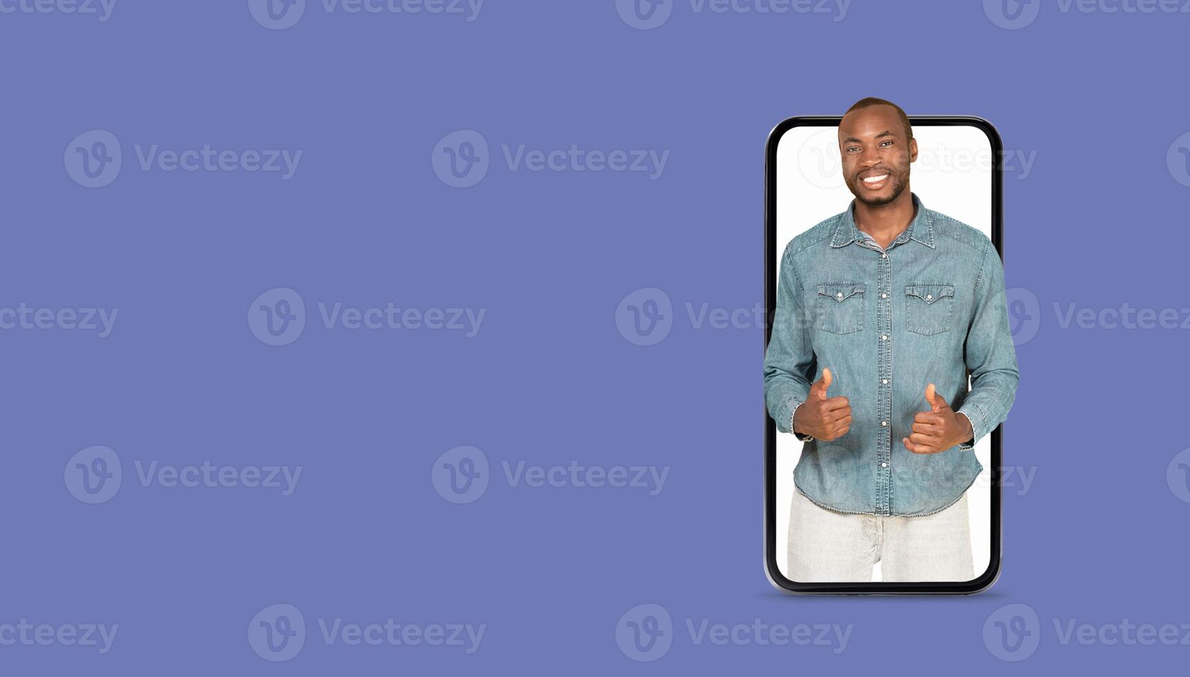 A young man smiles and gestures while holding a smartphone with a large screen. He seems to be using an application for social media or communication against a plain background. photo