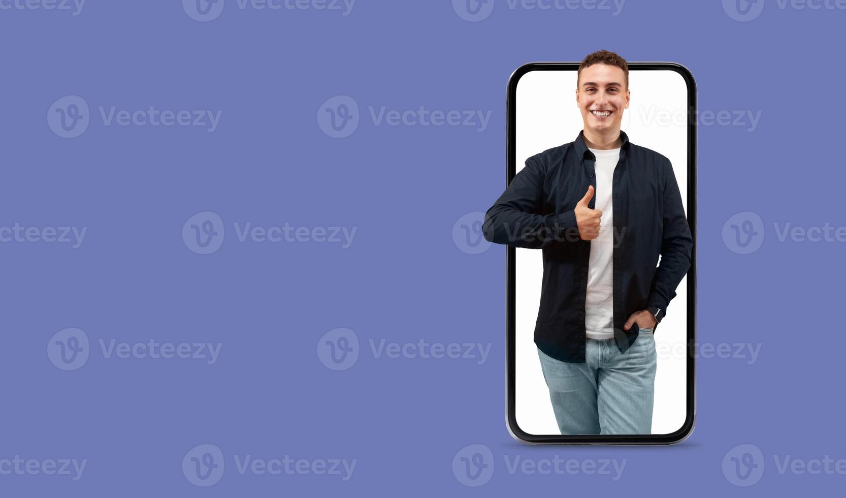 A young man stands confidently, smiling and giving a thumbs up gesture. He holds a smartphone with a large screen, symbolizing communication and connection in modern technology. photo