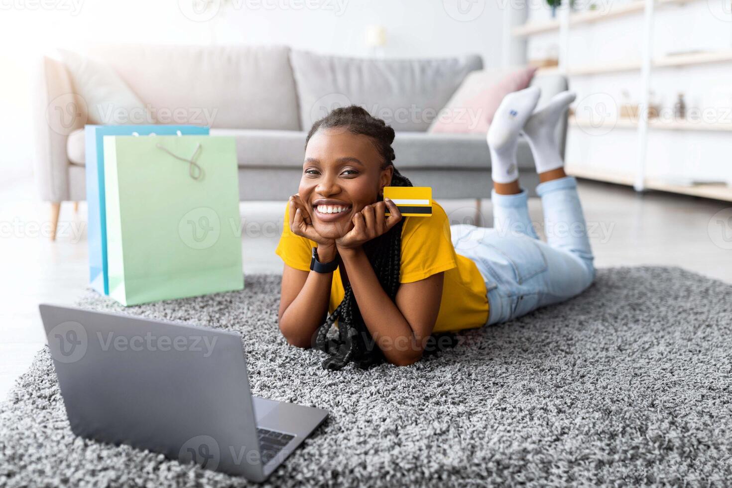 Shopaholism and e-commerce. Millennial Afro lady using credit card for remote payment, shopping online via laptop, purchasing goods in web store, lying on floor with gift bags at home photo