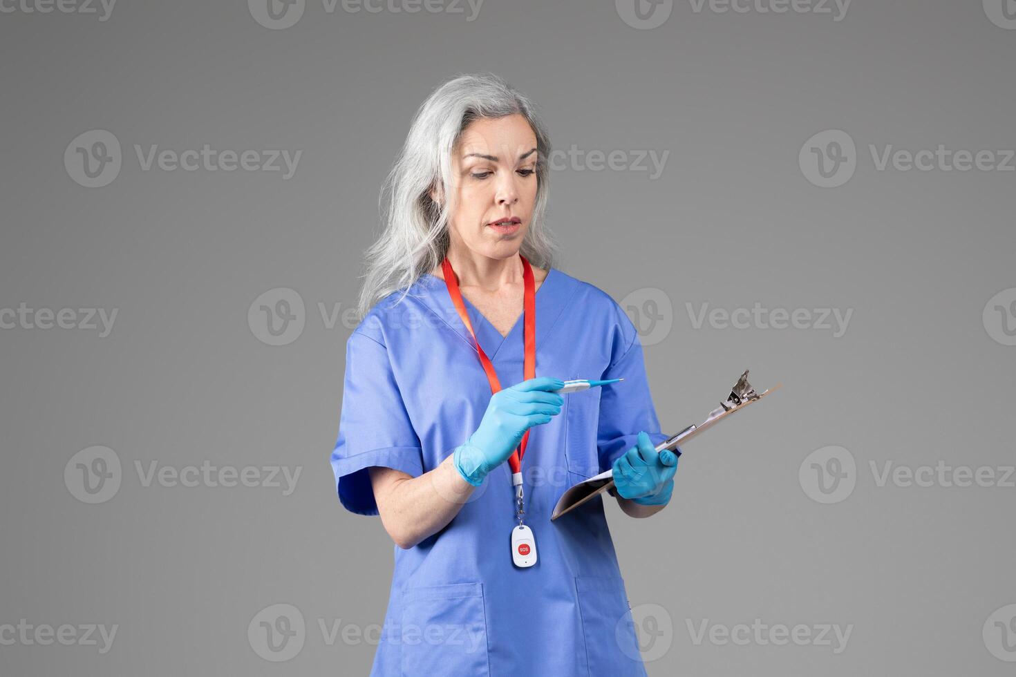 A mature woman dressed in blue scrubs and gloves holds a clipboard while writing with a pen. She appears focused and engaged in her healthcare duties in a neutral, isolated background. photo