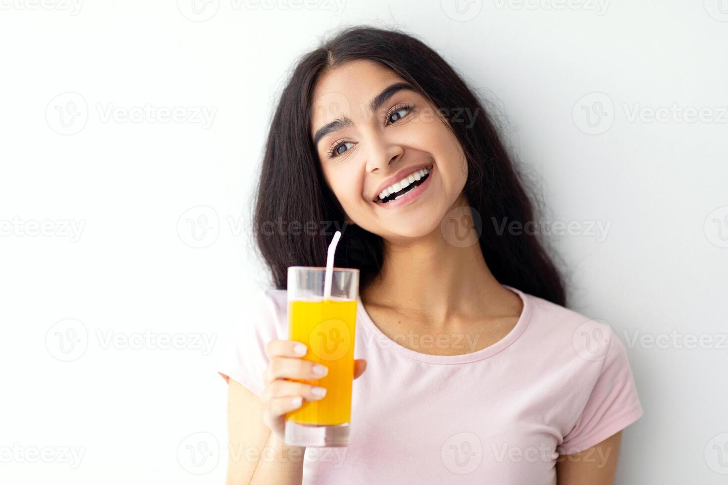 Summer detox and healthy eating concept. Smiling Indian woman with glass of fresh orange juice against white wall. Positive Eastern lady enjoying her delicious fruit drink photo
