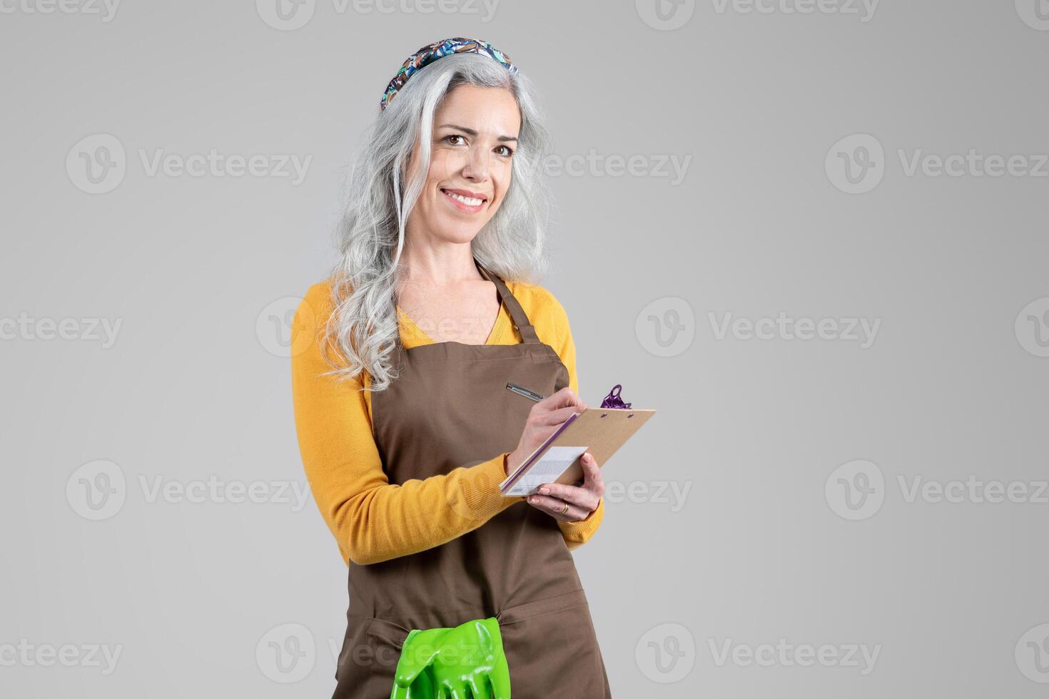 A middle aged woman stands confidently in her gardening attire, smiling as she holds a clipboard. Her long, gray hair is styled, and she embodies a passion for gardening and farming, copy space photo