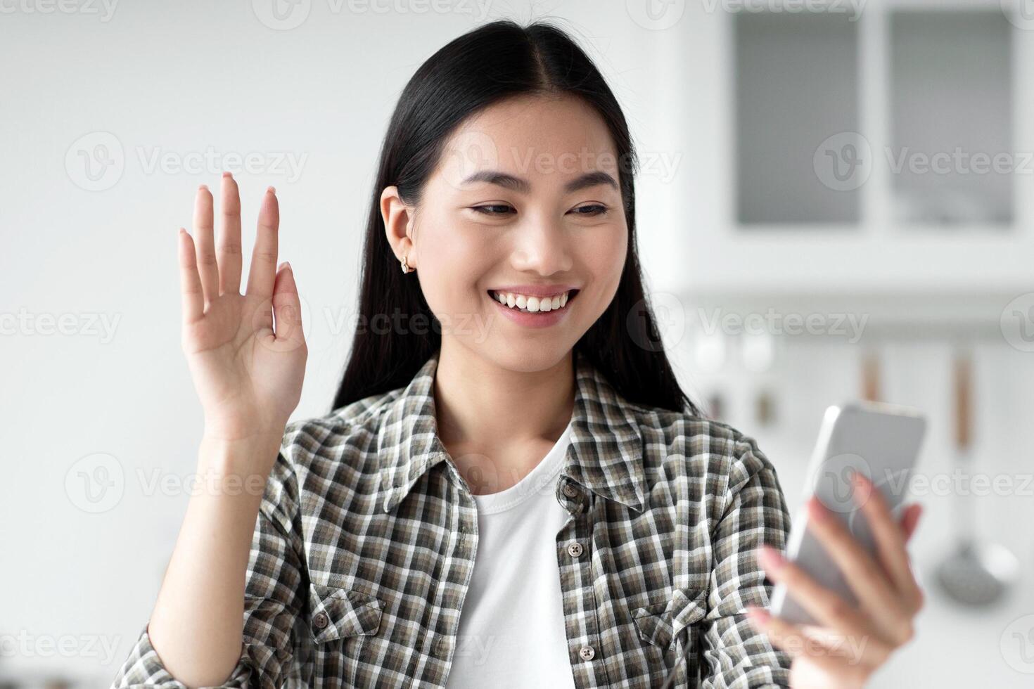 Happy young asian woman having chat with boyfriend during breakfast, kitchen interior, copy space. Positive korean lady student sitting at table, eating healthy food, waving at smartphone screen photo