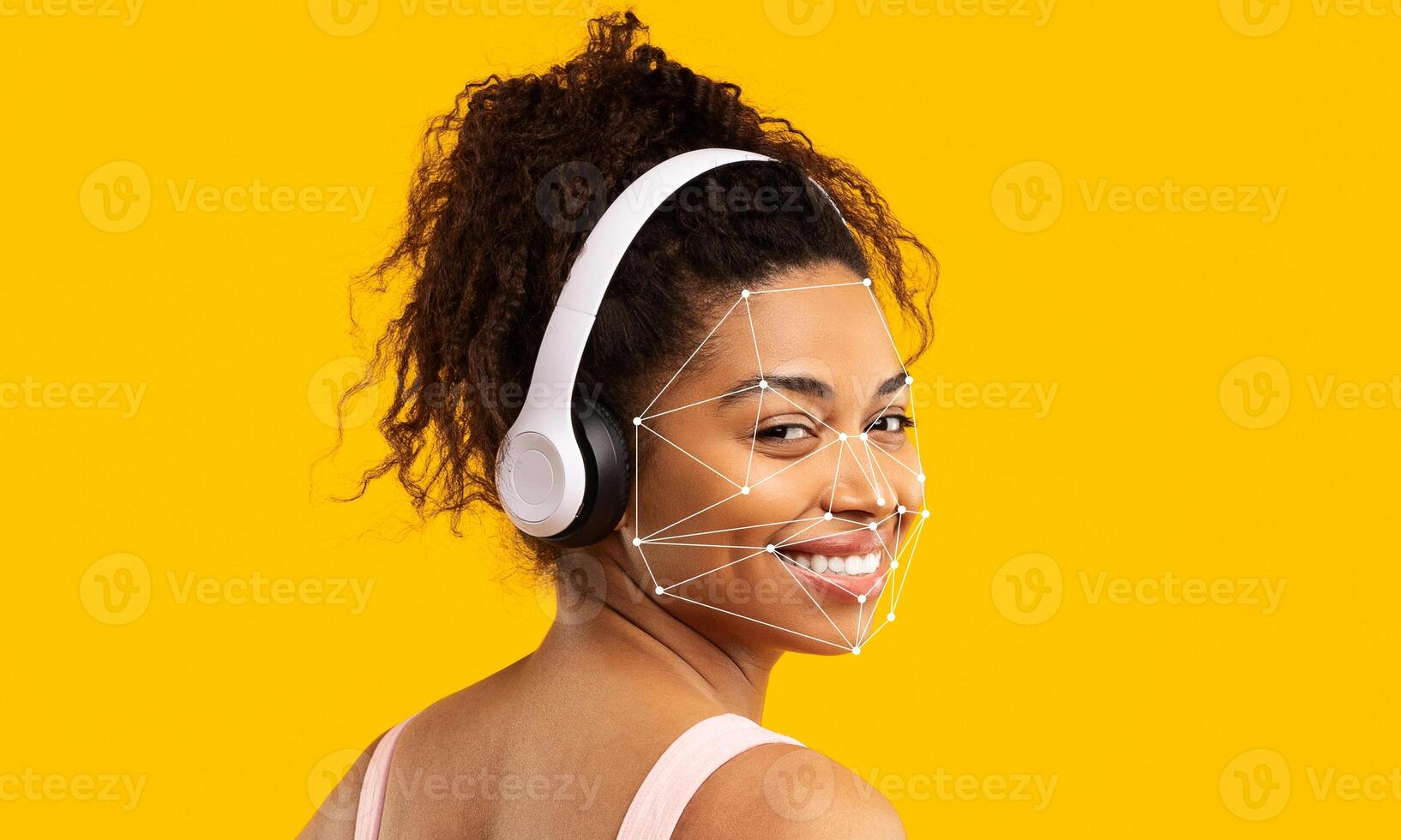 A woman with curly hair wears headphones while smiling. Facial recognition technology is analyzing her features against a bright yellow background. The moment captures innovation in identification. photo