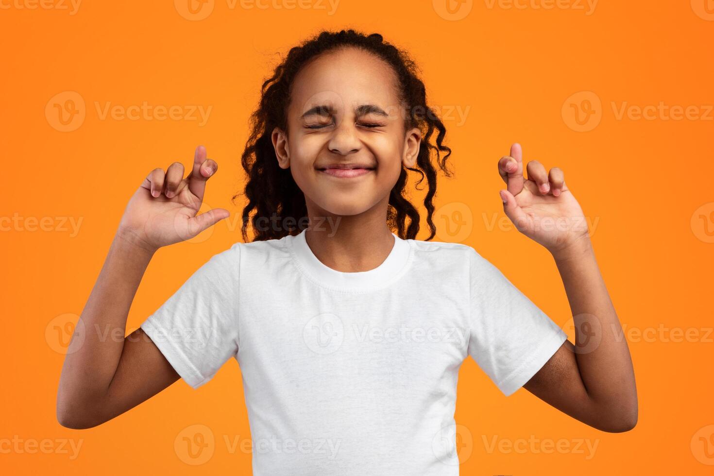 Luck And Superstition Concept. Cheerful black teen gesturing and crossing fingers in hope, anticipating something with faithful expressions and closed eyes, isolated on yellow studio background photo