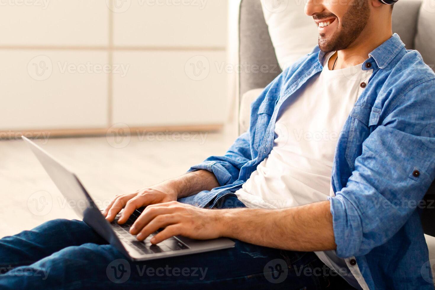Successful Career And Distance Job Concept. Portrait Of Happy Jewish Guy Working On Laptop Wearing Wireless Headset Sitting On Floor Leaning On Sofa At Home Office, Modern Interior, Free Space photo