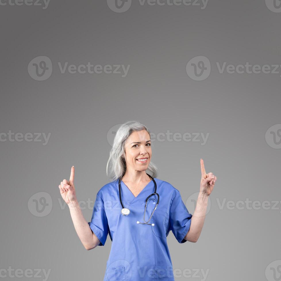A middle aged woman wearing scrubs and a stethoscope smiles while pointing upwards at copy space. The setting is isolated with a plain gray background, emphasizing her enthusiasm and professionalism. photo