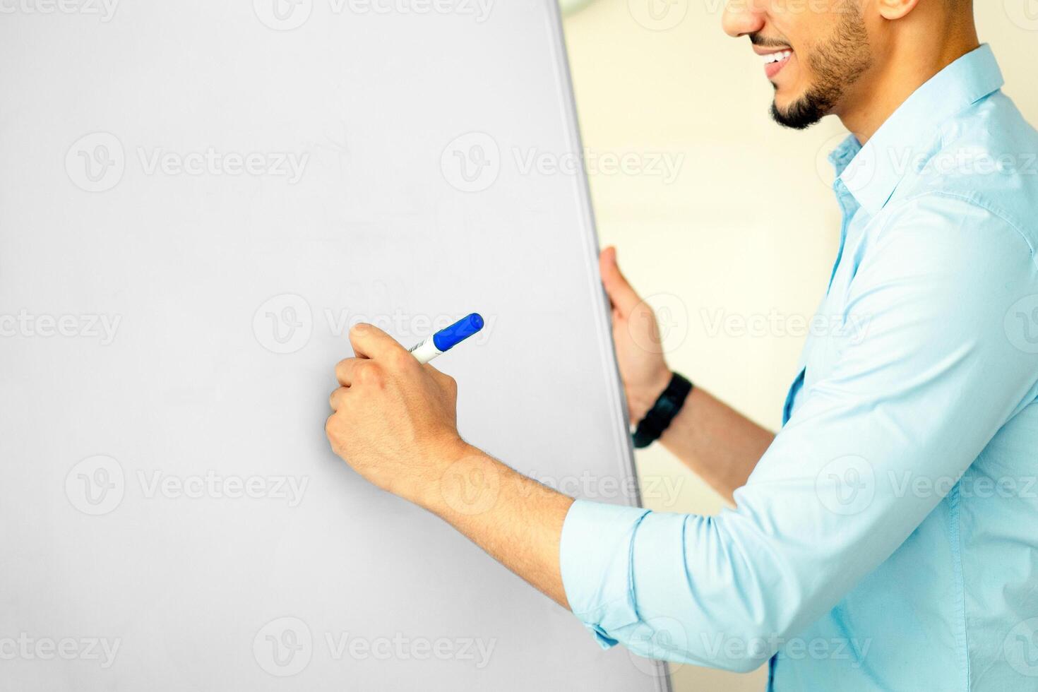 Distance learning concept. Happy Arab guy tutor giving online English class, writing on blackboard at home office. Positive school teacher explaining rules to students on web photo