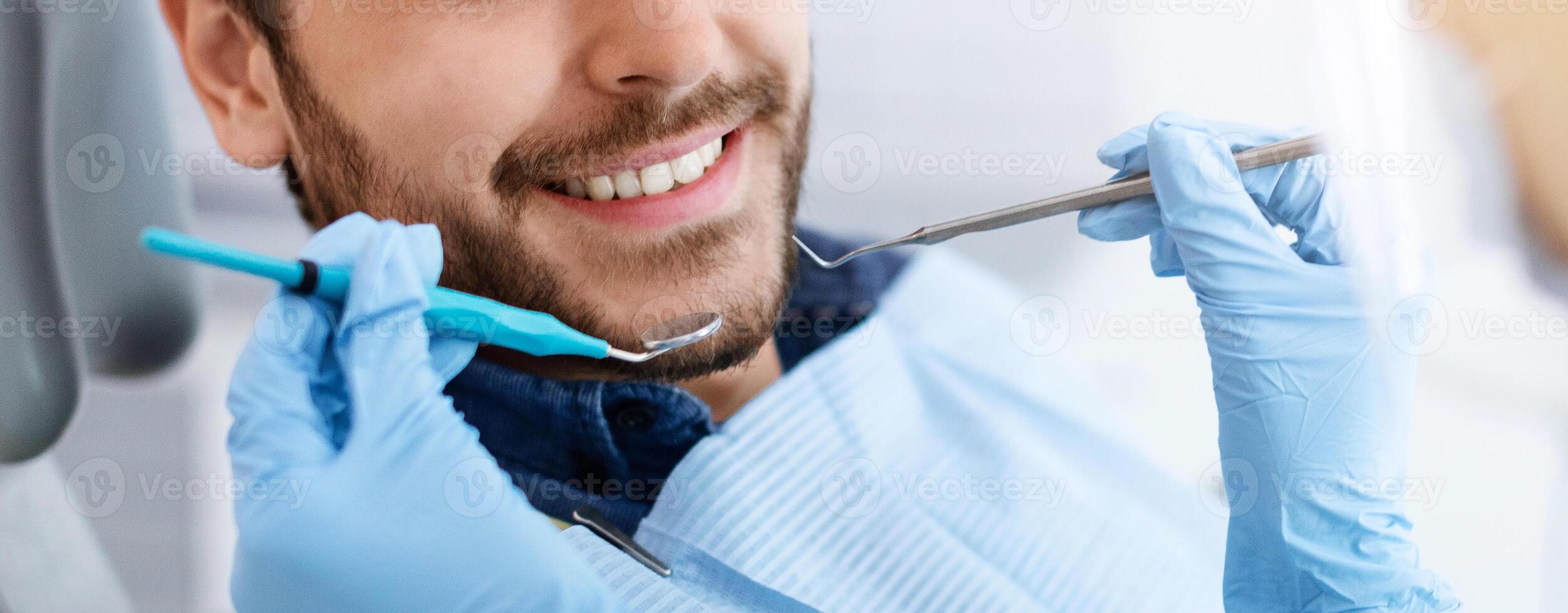 Closeup of dental clinic patient smile and dentist hands holding tools, unrecognizable cheerful bearded man attending dentistry, panorama. Banner for modern dental clinic advertisement photo