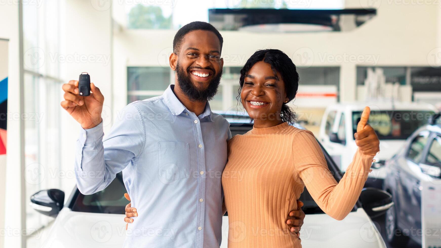 Happy Black Couple Holding Keys From New Car And Showing Thumb Up, Cheerful Young African American Spouses Purchased Automobile In Dealer Showroom, Looking And Smiling At Camera, Free Space photo