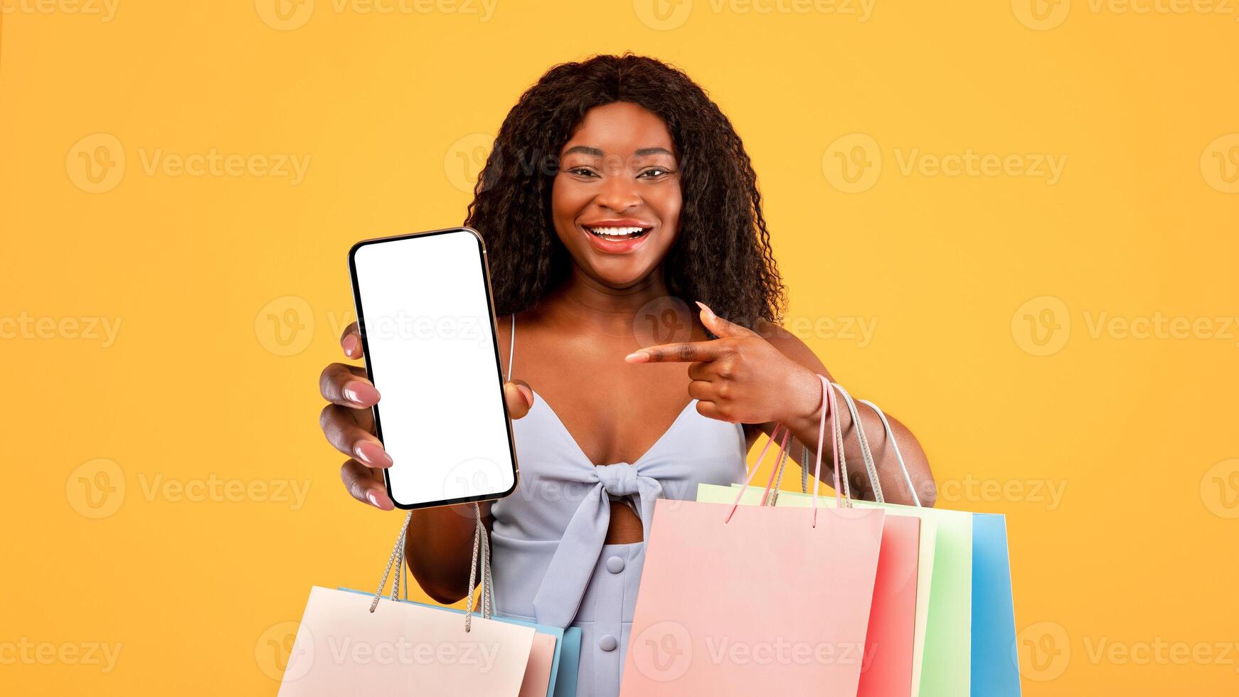 Smiling African American woman with shopper bags pointing at cellphone with empty screen, advertising new mobile shopping app over orange studio background, mockup for your design photo