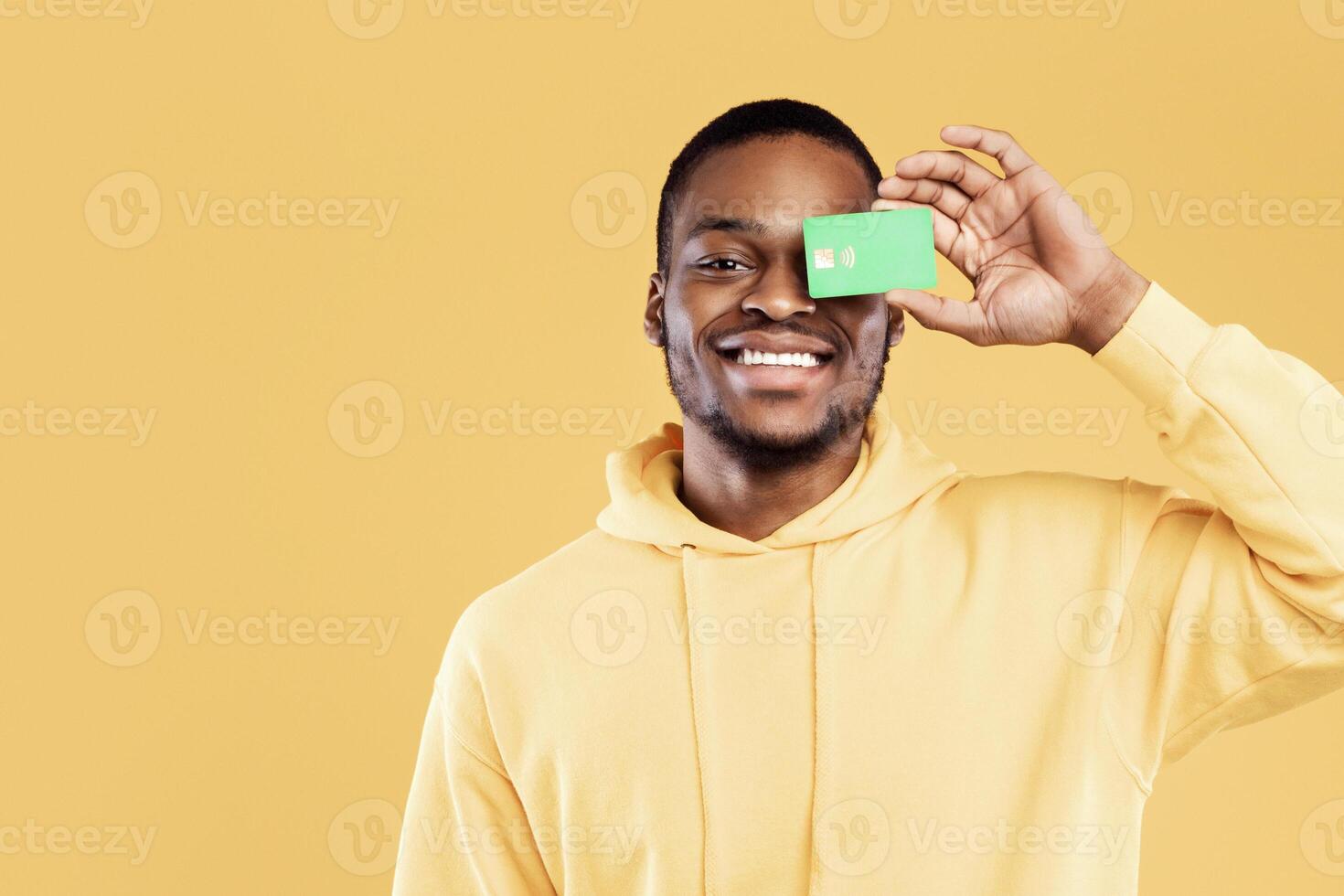 Positive Black Guy Posing With Credit Card Covering His Eye Standing Over Yellow Studio Background, Smiling To Camera. Finances, Bank Offer Concept. Panorama photo