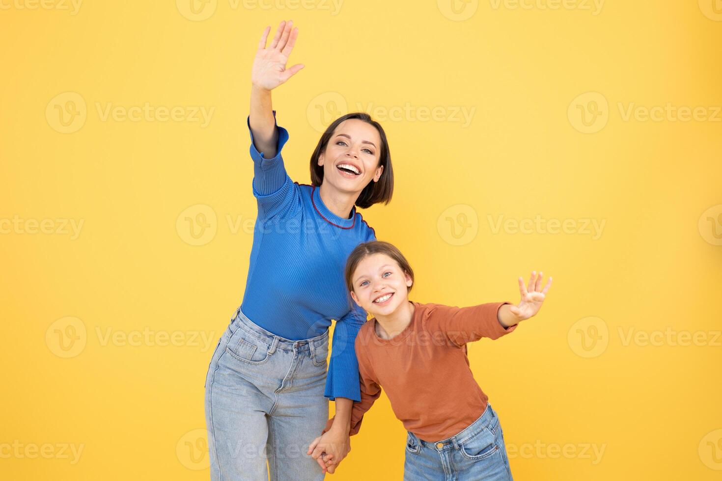A mother and her preteen daughter are smiling and waving happily. They are holding hands, expressing affection and love in a cheerful setting with a vibrant yellow backdrop. photo