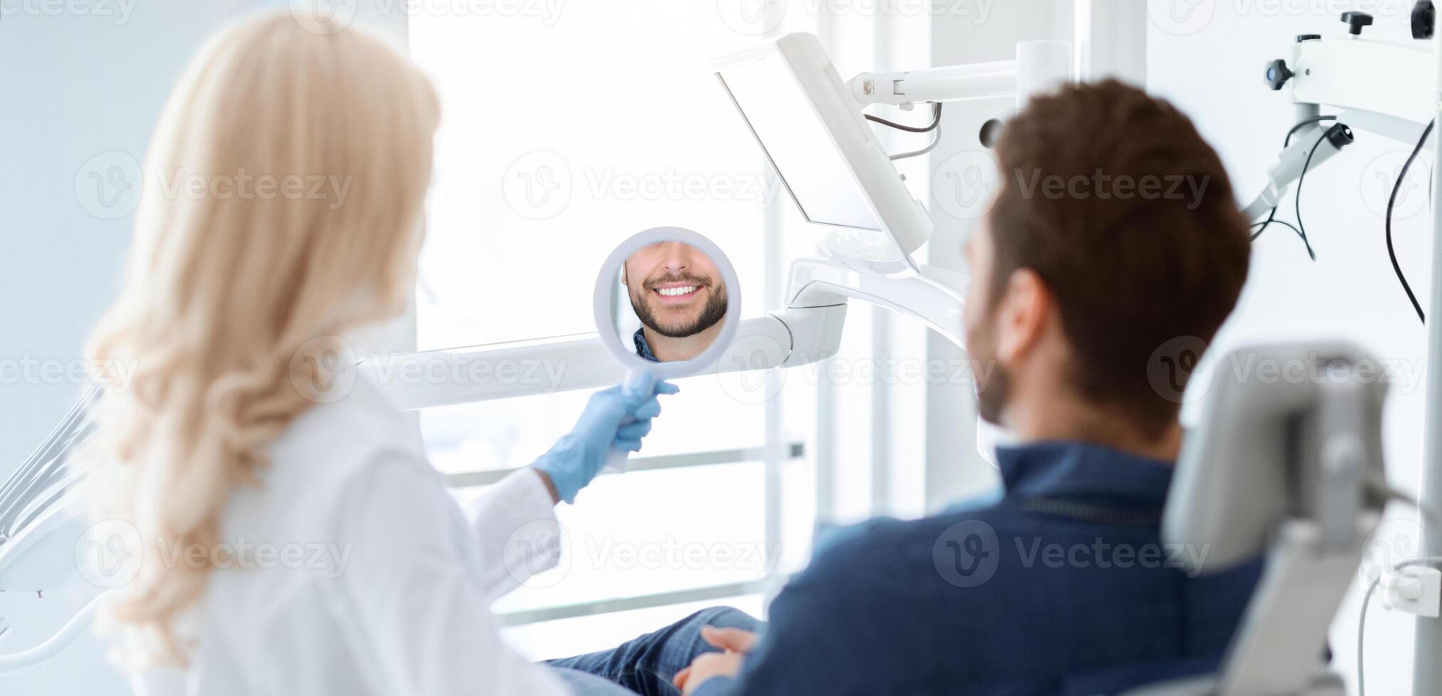 Female dentist holding mirror for smiling bearded man patient, stomatologist and happy man checking results of treatment, healthy white smile reflection in small dental mirror, back view photo