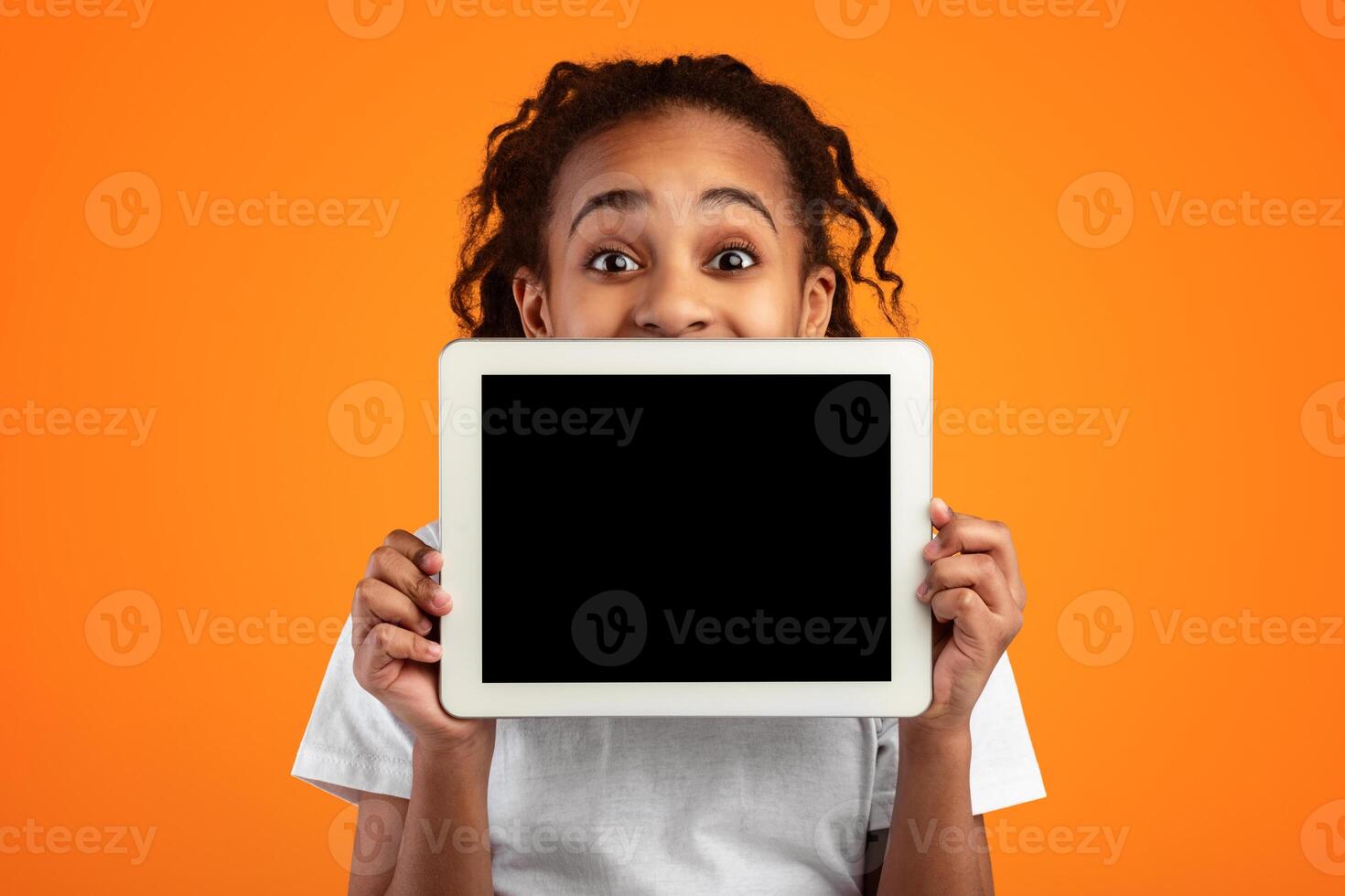 Great Application. Excited Black Girl Holding And Showing Digital Tablet With Empty Screen For Mockup, Covering Face And Mouth, Recommending New App Posing Standing Over Yellow Studio Background photo