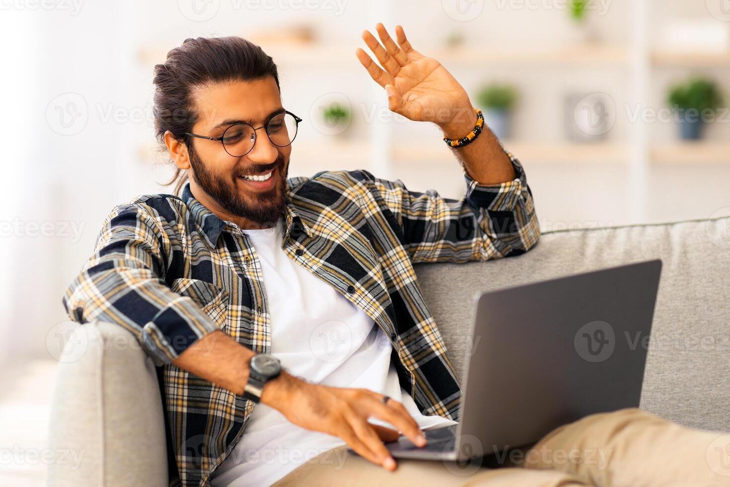 Cheerful long-haired, bearded indian guy sitting on couch in living room, using laptop, having conference with client or girlfriend from home, smiling and waving at notebook screen, copy space photo