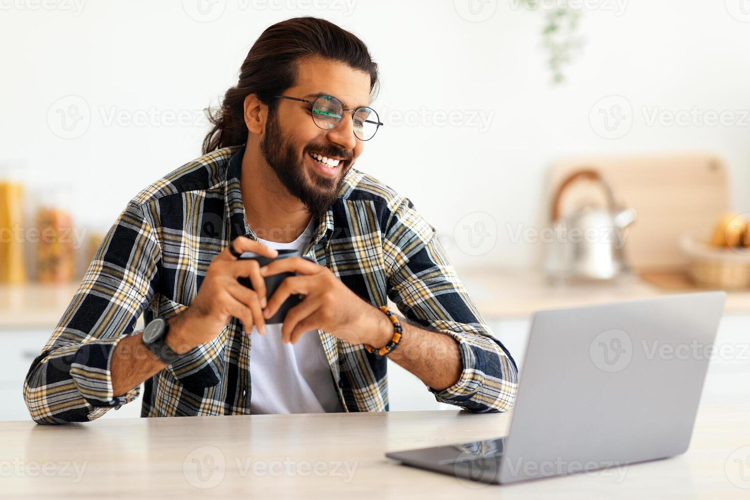 Cheerful indian guy with glasses looking for job online, sitting at table, using laptop, drinking coffee, kitchen interior, copy space. Happy arab man student working online from home, remote job photo