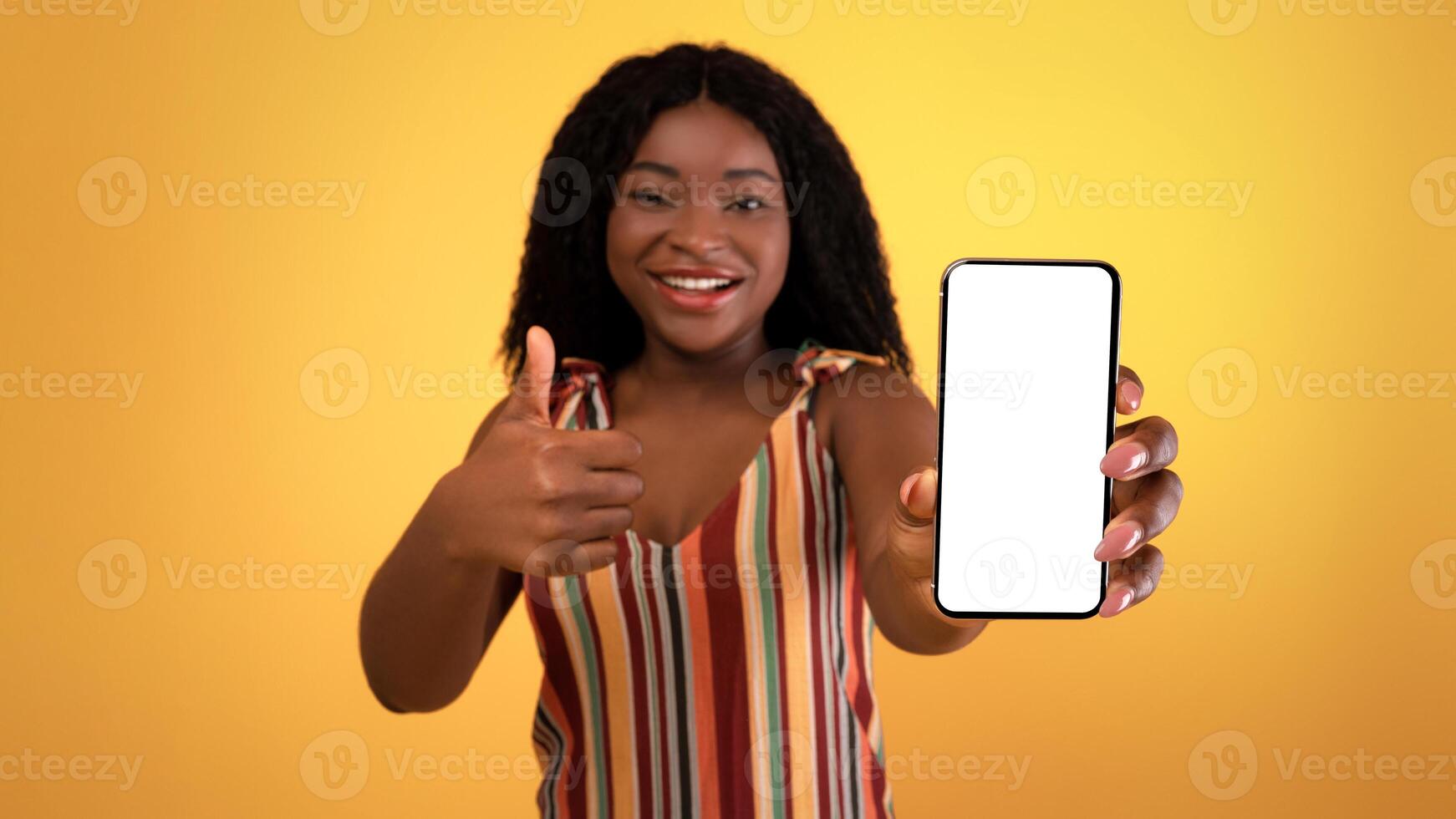 Lovely black woman demonstrating cellphone with empty black screen, showing thumb up, recommending new cool mobile shopping app on orange studio background, mockup for design photo