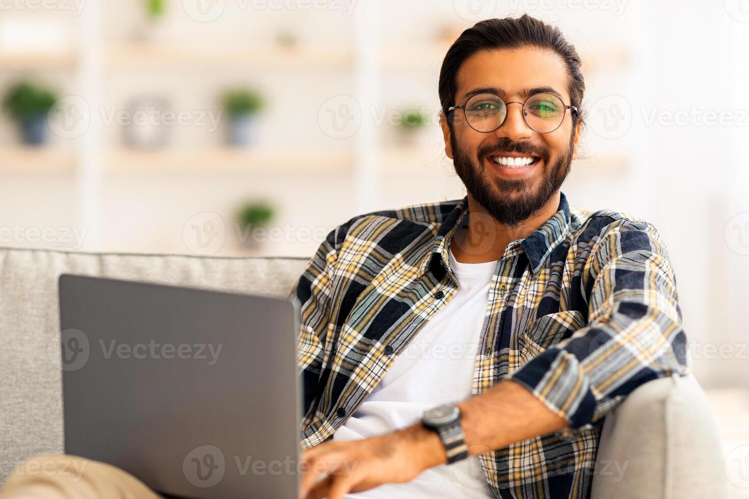 Positive indian guy reclining on couch with laptop, watching movie or chatting with ladies on dating website while relaxing at weekend, middle-eastern freelancer working from home, copy space photo