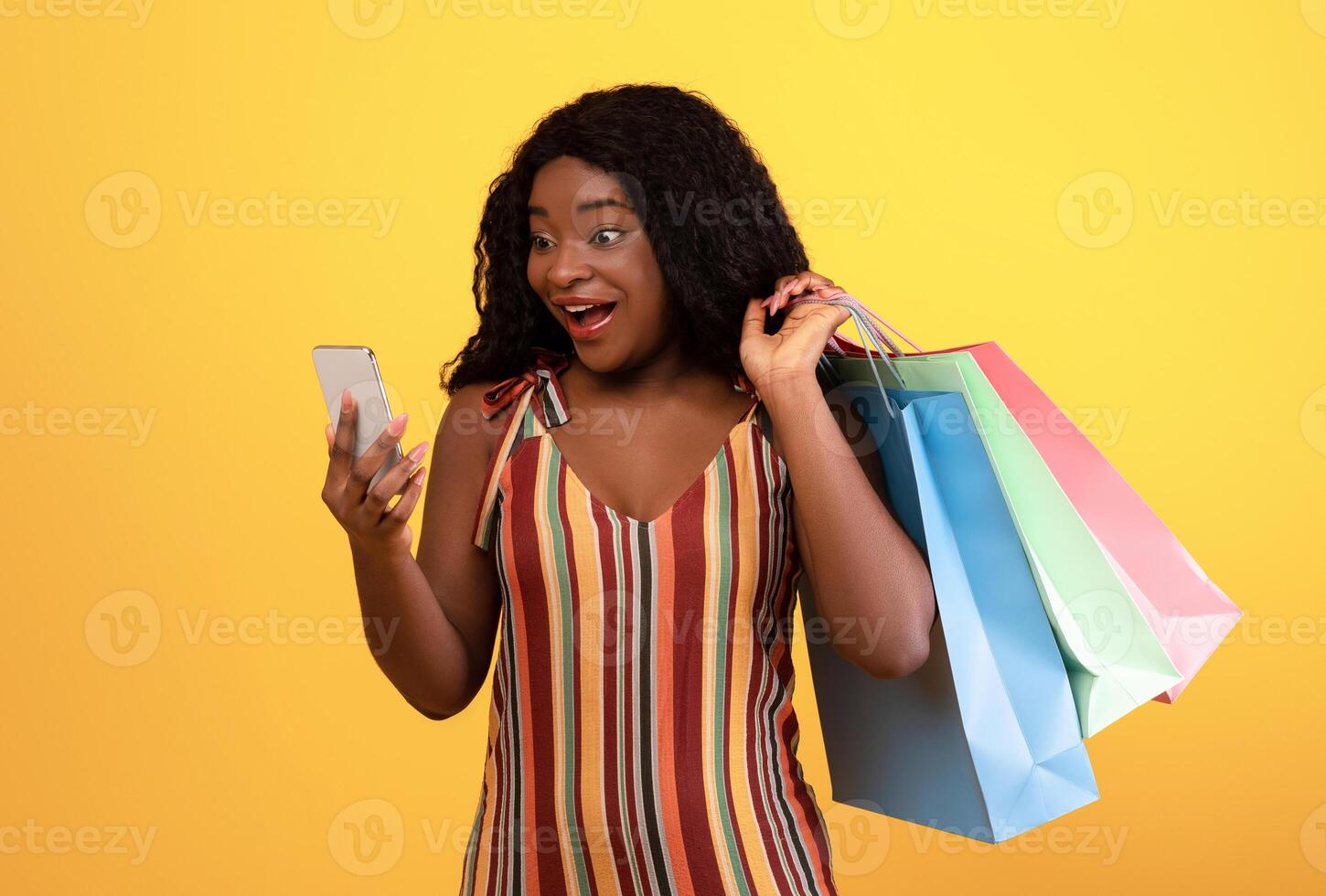 Huge sale in web store. Shocked black lady with shopping bags looking at smartphone, using new mobile app, purchasing things online over orange studio background photo