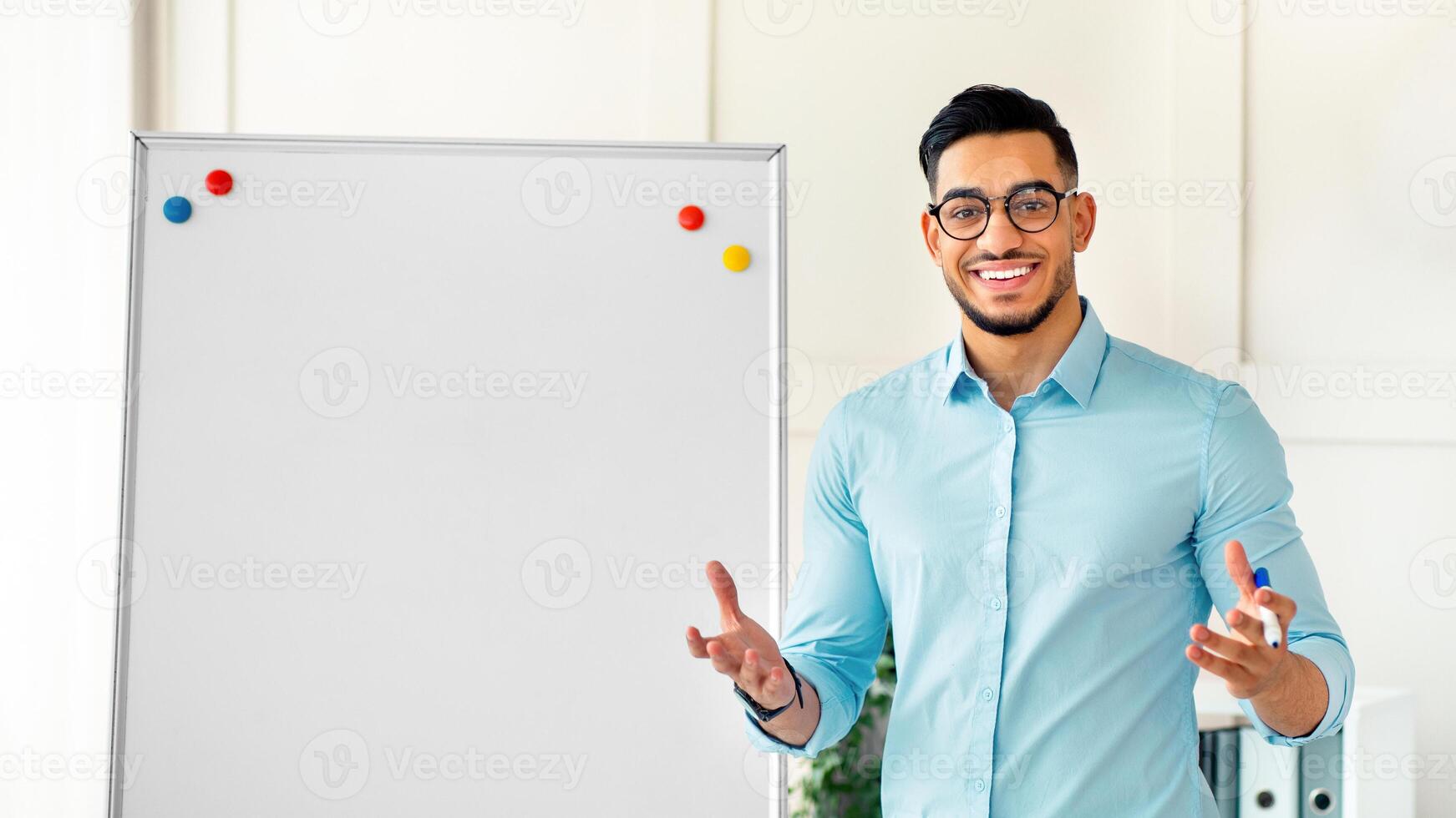 Smiling Arab guy teacher giving remote class, standing near blackboard, explaining English rules to pupils from home office. Online tutoring and distance education during covid lockdown concept photo