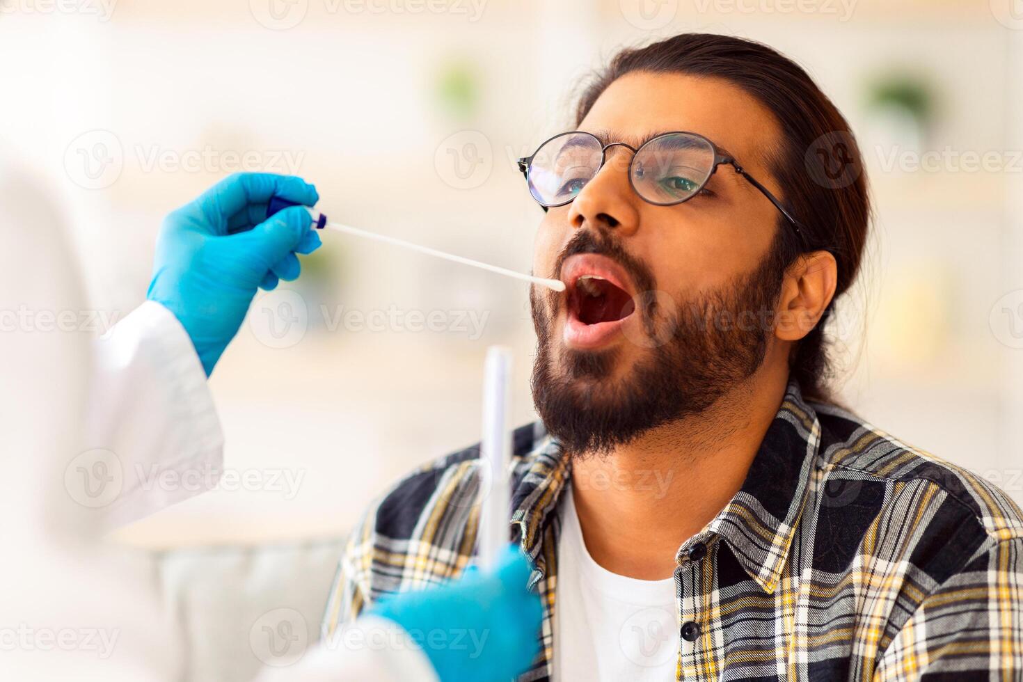 Indian guy getting coronavirus swab test from throat at home, unrecognizable nurse in protective medical costume making covid-19 swab test for sick patient young man at home, closeup photo