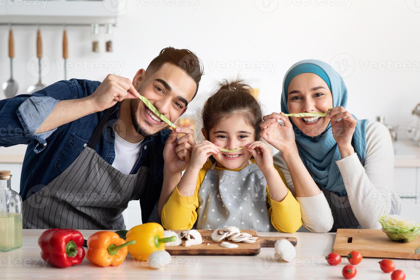 Portrait Of Joyful Muslim Family Of Three Fooling Together In Kitchen, Making Mustaches With Lettuce Slices And Smiling At Camera, Cheerful Arabic Parents And Little Daughter Having Fun At Home photo