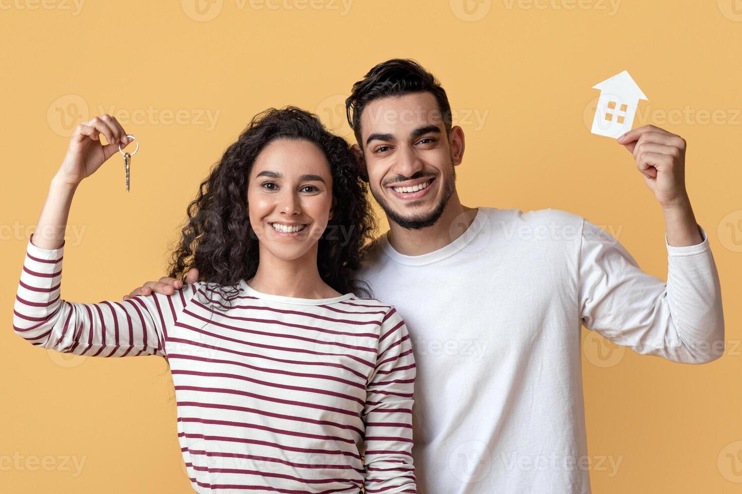 Family Housing. Happy Arab Couple Holding Home Keys And Paper House Icon, Joyful Middle Eastern Spouses Celebrating Moving To Their Own Apartment, Standing Over Yellow Studio Background, Free Space photo