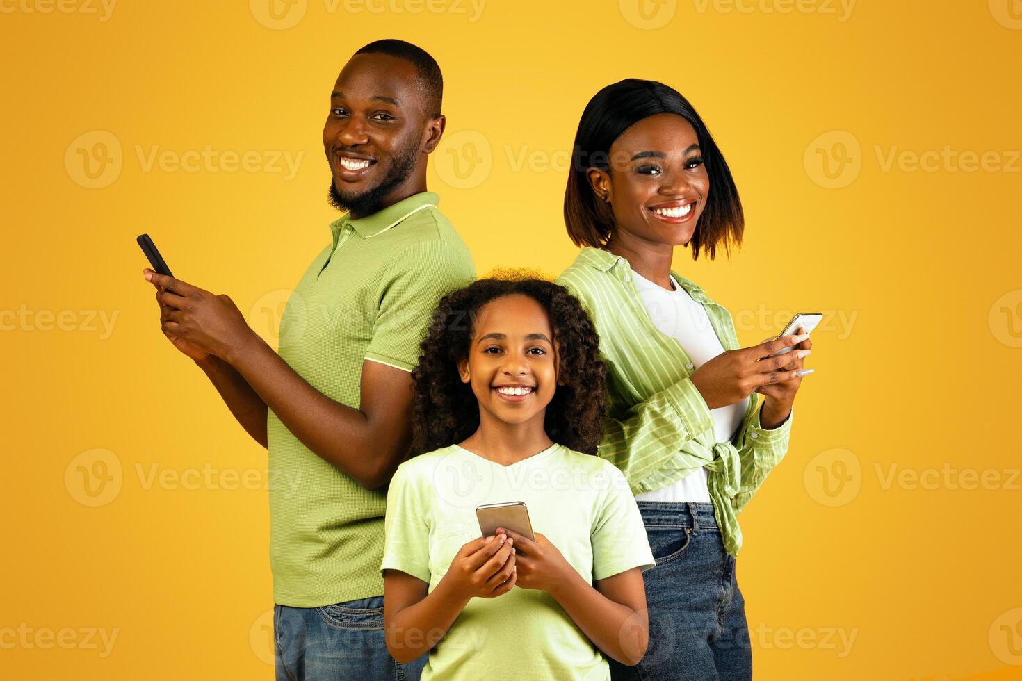 Modern family. Excited african american father, mother and daughter using smartphones while standing over yellow studio background and smiling to camera photo