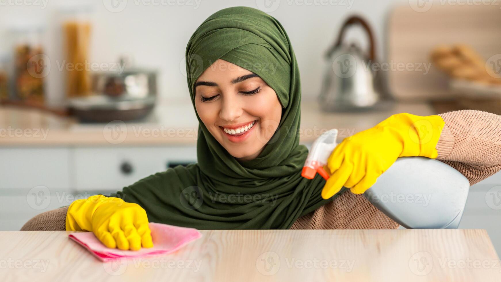 Closeup portrait of happy islamic housewife in hijab and protecitve yellow rubber gloves cleaning dining table at kitchen. Positive maid keeping her home tidy, wiping surface with microfiber cloth photo