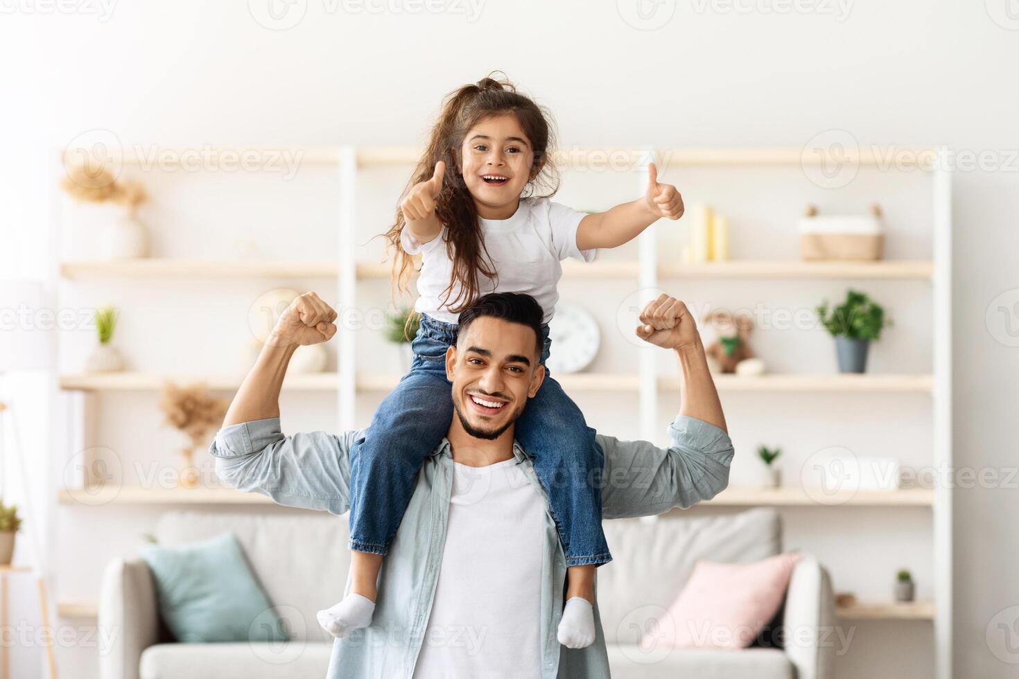 Playful middle-eastern father and daughter having fun at home, cheerfully posing at camera, copy space. Happy arab man dad carrying little girl kid on his shoulders and showing strong muscles photo