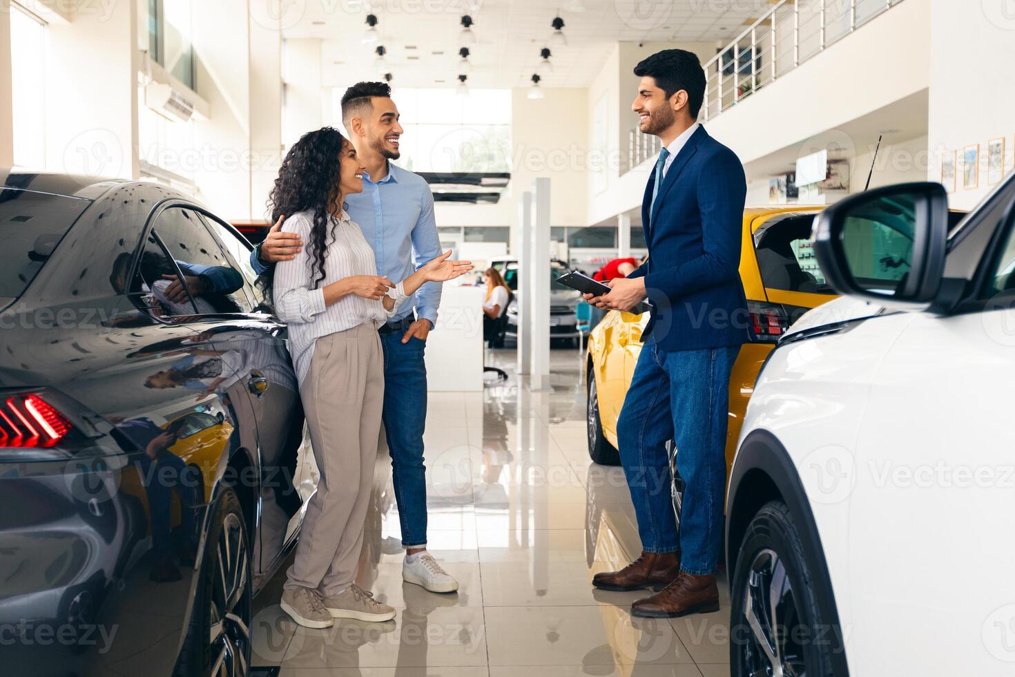 Wealthy middle eastern young couple having conversation with good-looking sales assistant at luxury auto showroom, looking for new car, gesturing and smiling, full length photo, panorama photo