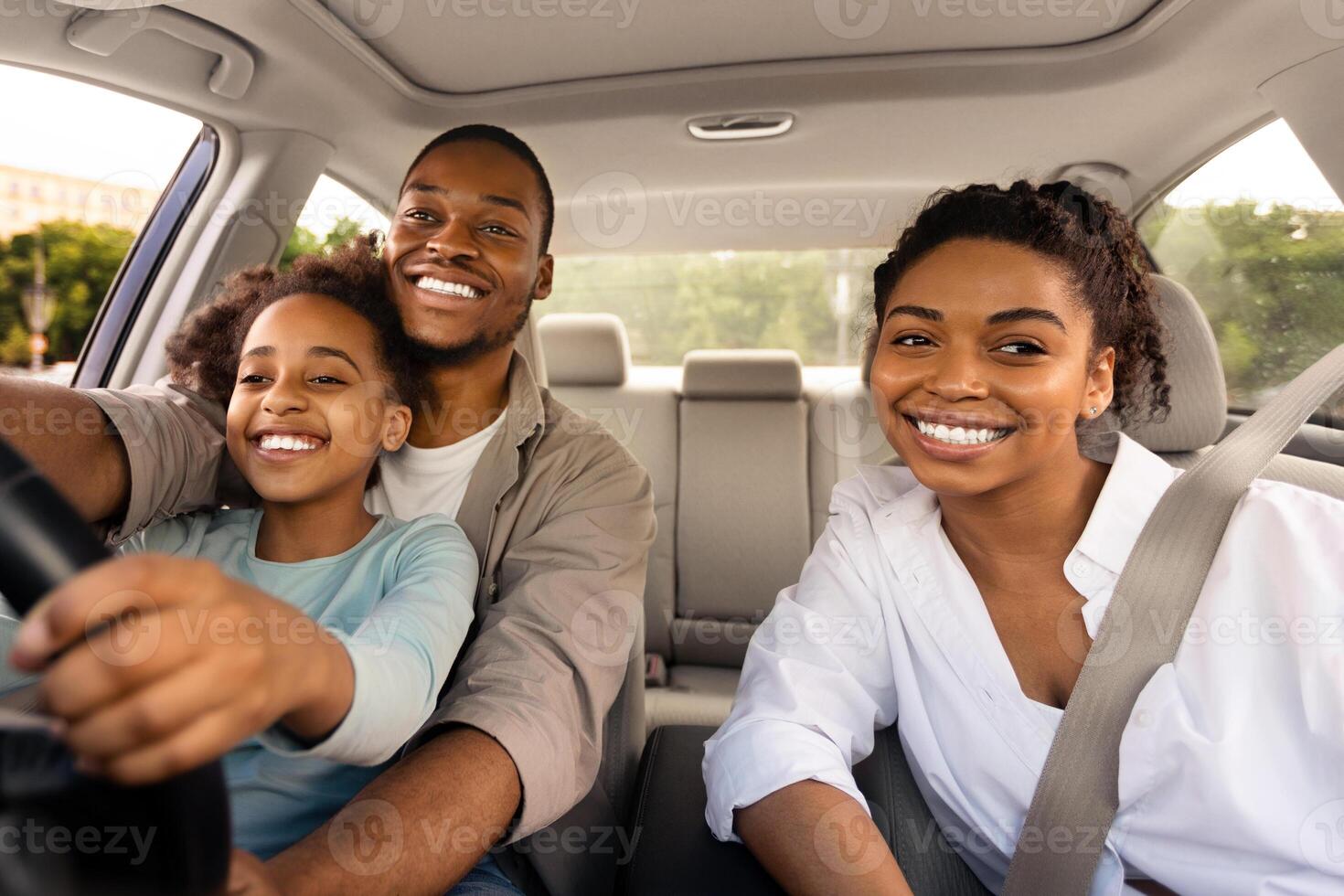 Happy Black Family Driving Car Together, Preteen Daughter Learning To Drive Sitting On Father's Knees Having Fun Riding In City. Parents And Kid Having Road Trip Adventure Concept photo