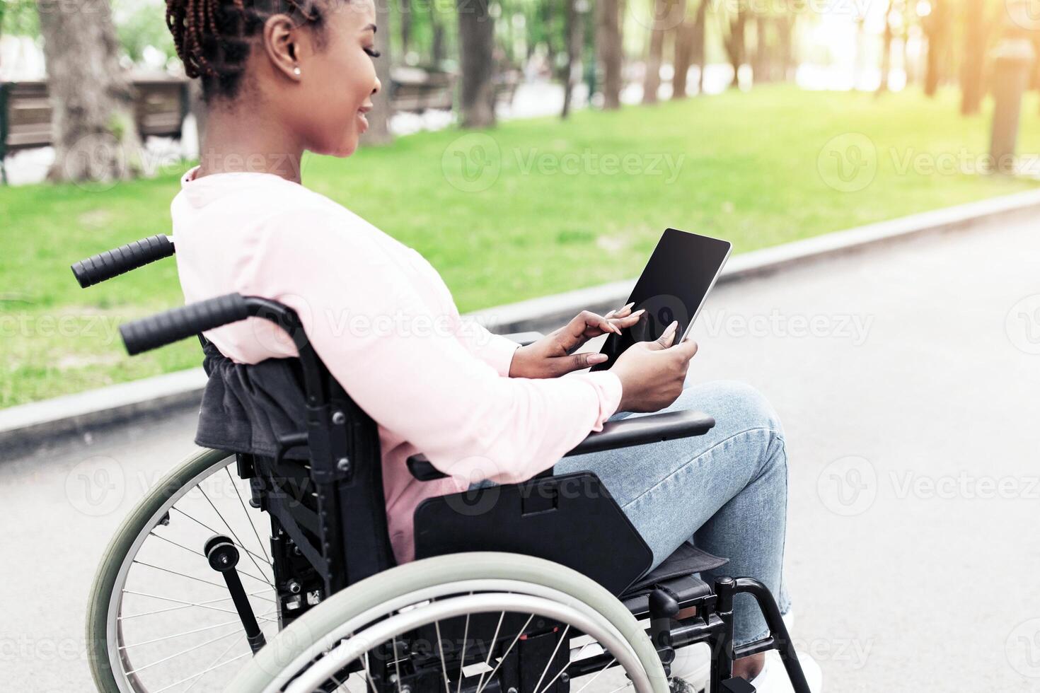 Young black woman in wheelchair using tablet computer with empty screen at city park, mockup for app or website. Millennial African American lady with disability working on touch pad outdoors photo