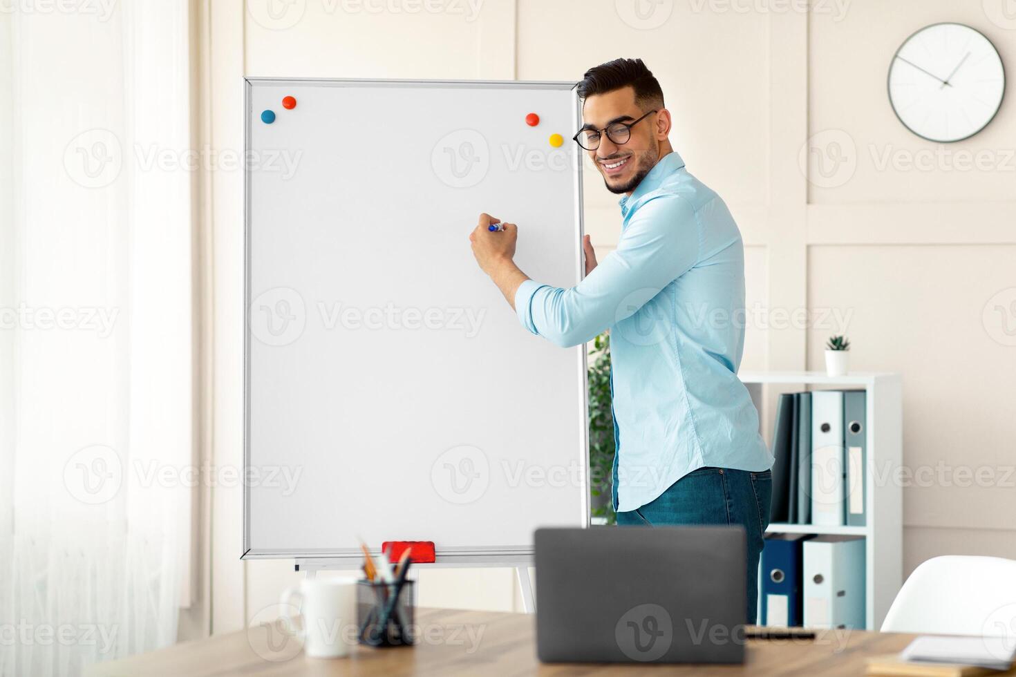 Portrait of positive Arab guy teacher giving online English lesson, writing grammar rules on blackboard, speaking to students on webcam. Distance education during covid outbreak photo