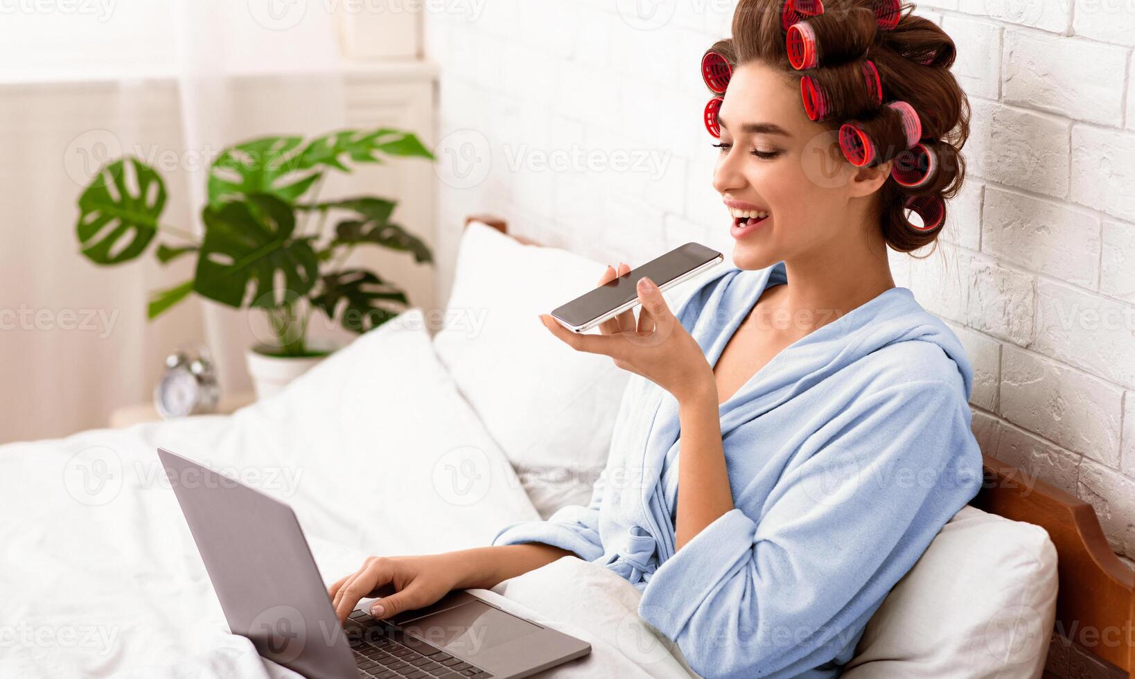 Cheerful Lady Using Voice Assistant On Cellphone Working At Laptop Computer Sitting In Bedroom, Curling Hair With Curlers. Distant Work From Home, Freelance, Smart Phone App Concept. photo