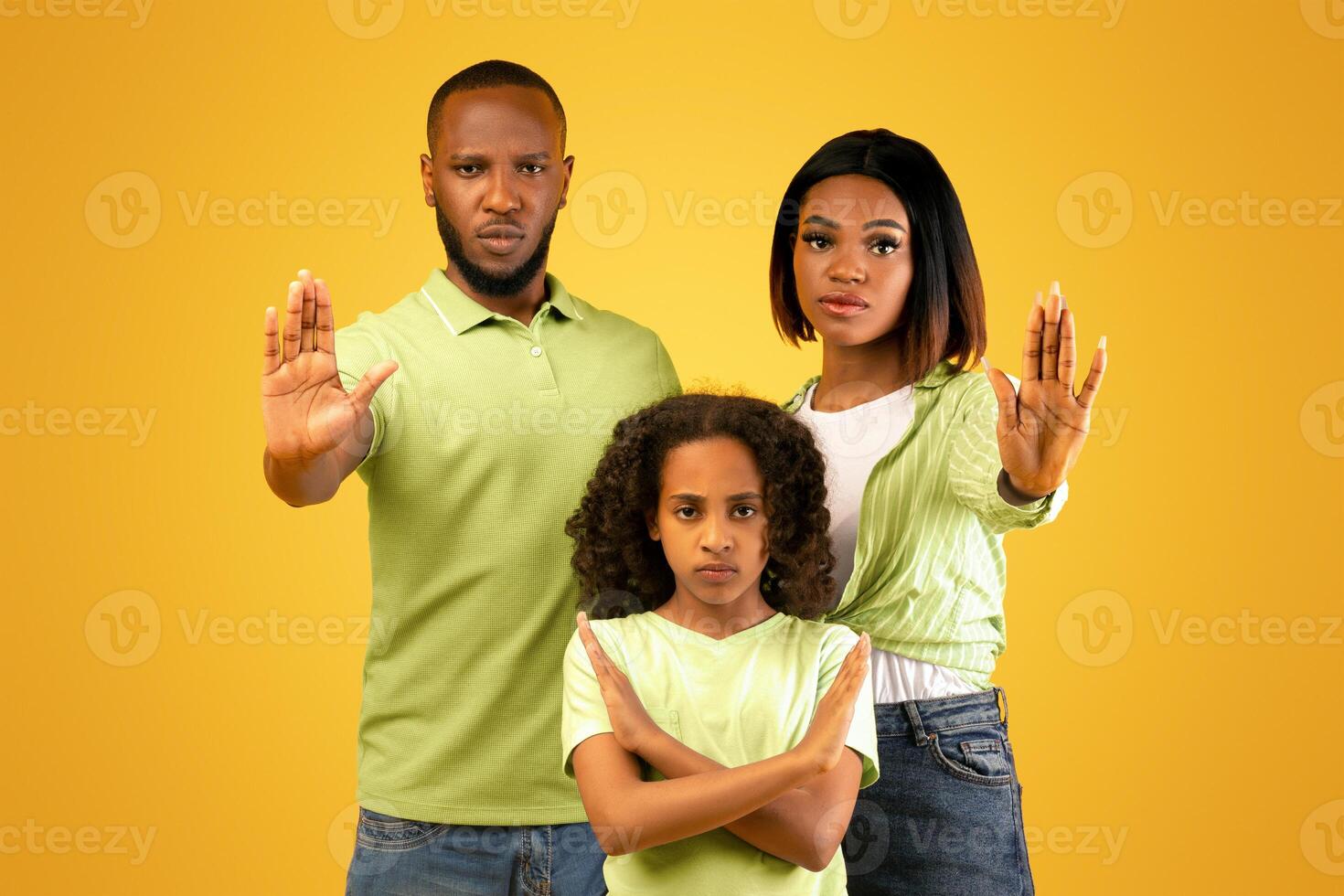 Protest, disagree and denial gesture. African american parents and daughter showing stop sign and girl crossed arms, standing on yellow background, studio shot photo
