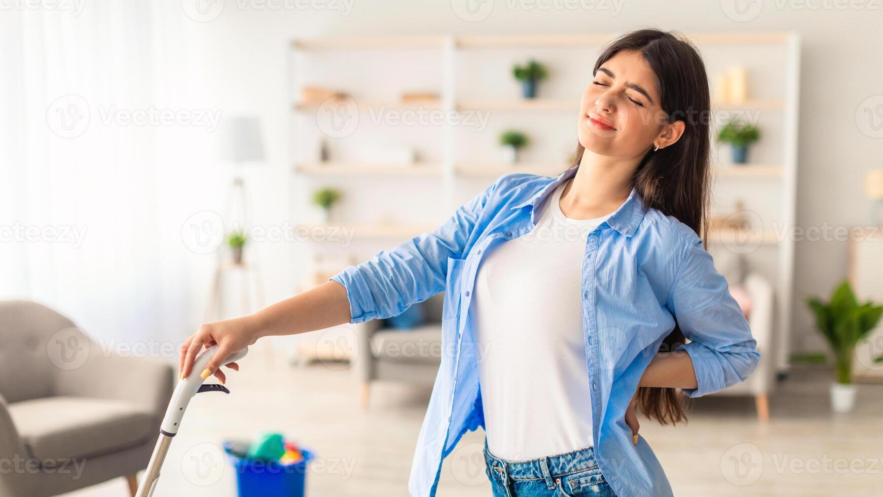Young woman having back ache from cleaning in her apartment, vacuuming floor, holding mop, free copy space. Tired millennial lady suffering from pain while cleaning dirty room photo